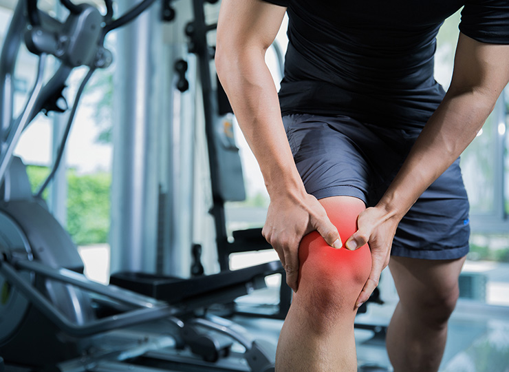 A close-up of a person in a gym holding their knee with both hands, with the front of the knee highlighted red to suggest pain or injury.
