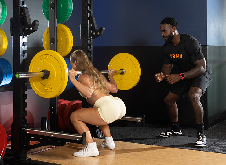Trainer coaching a woman during a barbell back squat in the gym.