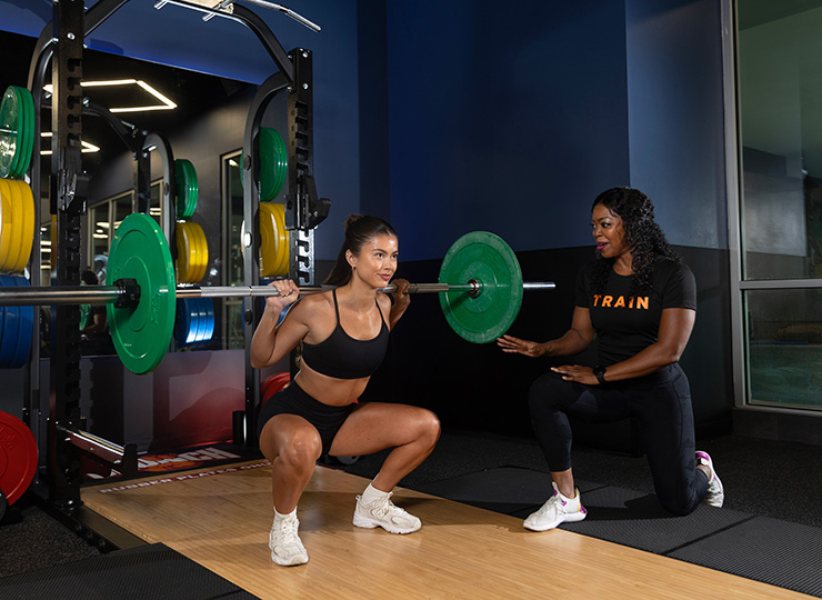 Woman doing a back squat while a trainer coaches beside her.