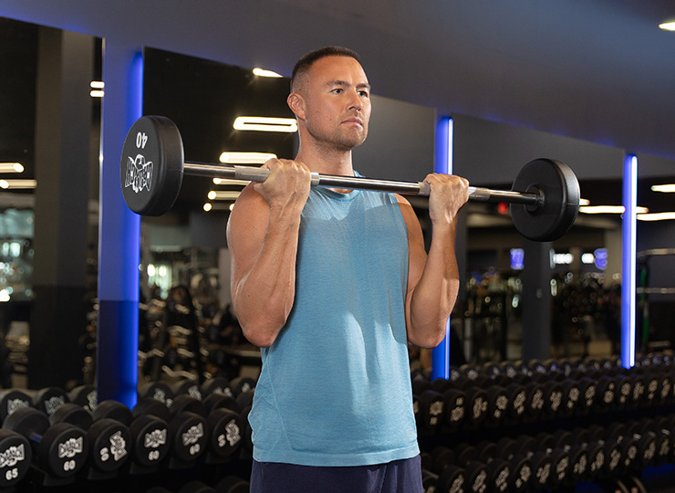 A man in a gym holds a barbell at shoulder height during a standing curl or front-rack position. He’s wearing a light blue sleeveless top, with rows of dumbbells and blue-lit mirrors in the background.