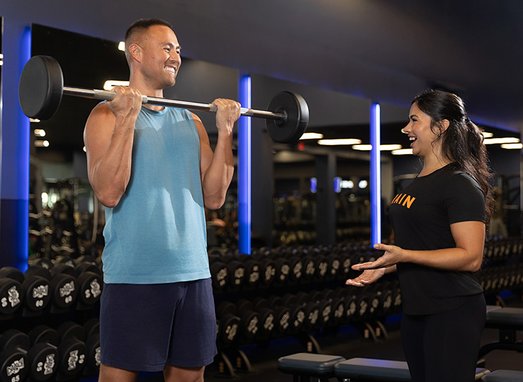 A man in a blue sleeveless shirt does a barbell curl in a gym while smiling at a woman standing beside him, who appears to be coaching or encouraging him. Rows of dumbbells and blue accent lights are visible in the background.