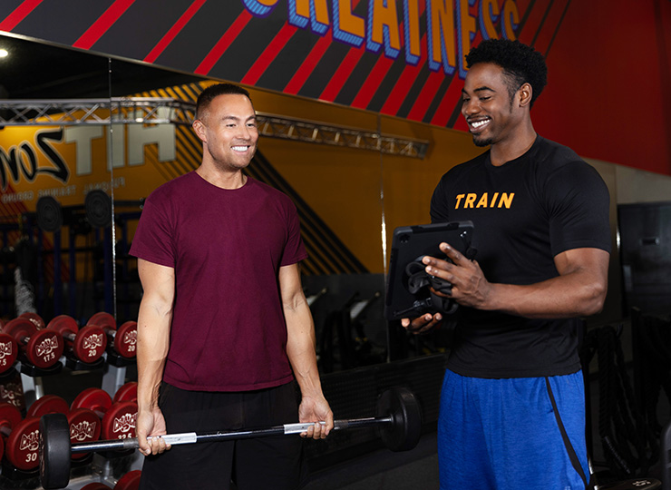 Screenshot of a website’s “Attachment details” page showing a personal trainer in a black “TRAIN” shirt holding a tablet while standing beside a gym member doing a barbell curl. They’re smiling in a weight area with red dumbbells and Crunch Fitness branding in the background, and an “Edit Image” button appears below the photo.
