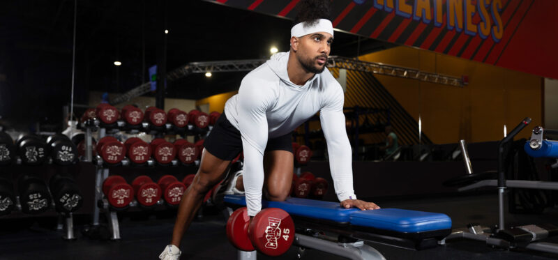 Wide banner image of a man in a gym doing a bench-supported row setup or core exercise position, wearing a white long-sleeve athletic top, black shorts, and a white headband. Red dumbbells and gym equipment are visible in the