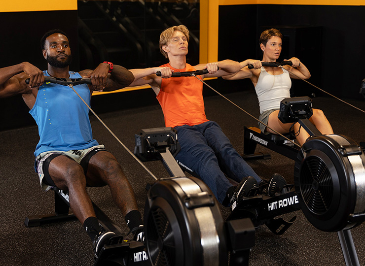 Three people are using rowing machines side by side in a gym, each pulling the handle toward their upper chest in the middle of a rowing stroke. The person on the left wears a blue tank top, the center person an orange sleeveless shirt, and the person on the right a light gray tank and shorts. The gym has black and yellow walls, and the image captures them in a synchronized, high-energy workout moment.