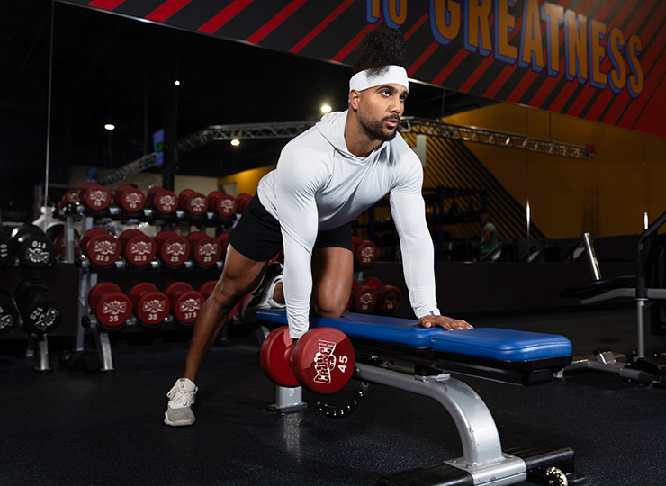 A man is doing a one-arm dumbbell row in a gym, bracing one hand and one knee on a blue bench while pulling a red dumbbell up with the other hand. He’s wearing a white long-sleeve hoodie, black shorts, and a white headband. Behind him are racks of red dumbbells and gym equipment, with the image focused on his upper-body form and workout stance.