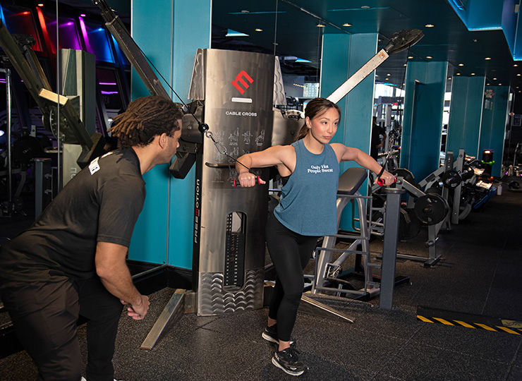 A woman is doing a cable chest fly in a gym while a trainer crouches nearby, watching her form. She’s in a staggered stance with arms extended out to the sides, holding the cable handles at chest height. The scene is set around a cable machine with blue walls, mirrors, and other gym equipment in the background.