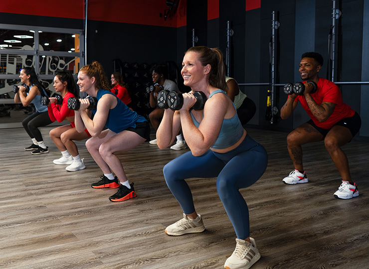 A group fitness class is doing goblet squats while holding dumbbells at chest height. Several participants are lined up in a studio with wood floors and dark walls, lowering into squat positions with focused expressions. In the foreground, a woman in a blue matching workout set is smiling while performing the movement, and other class members are visible behind her in colorful athletic wear.