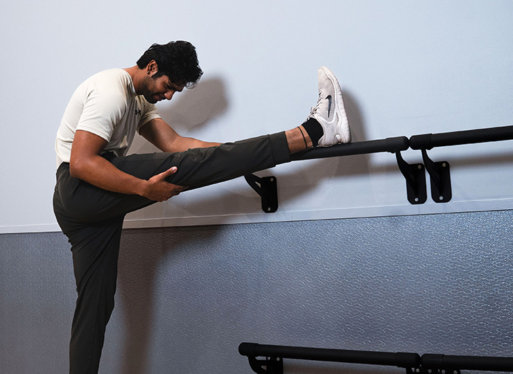 A man is stretching his hamstring by placing one leg up on a wall-mounted barre and leaning forward over it. He’s wearing a light athletic shirt, dark joggers, and white sneakers, and appears to be in a gym or studio space with a plain light wall and textured flooring.