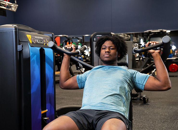 A man is using a seated shoulder press machine in a gym. He’s leaning back against the seat with both hands gripping the machine’s handles at shoulder height, preparing to press upward. He’s wearing a light blue athletic shirt, and other strength machines are visible in the background.