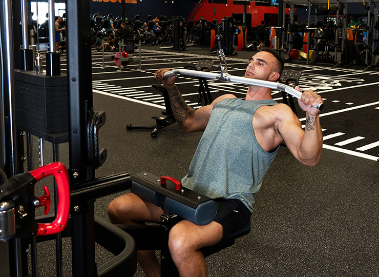A man is performing a lat pulldown on a cable machine in a gym. He’s seated with his thighs secured under the pads, leaning slightly back while pulling a wide bar down toward his upper chest. He’s wearing a light green sleeveless tank and black shorts, and the background shows a functional training area with turf-style flooring and gym equipment.
