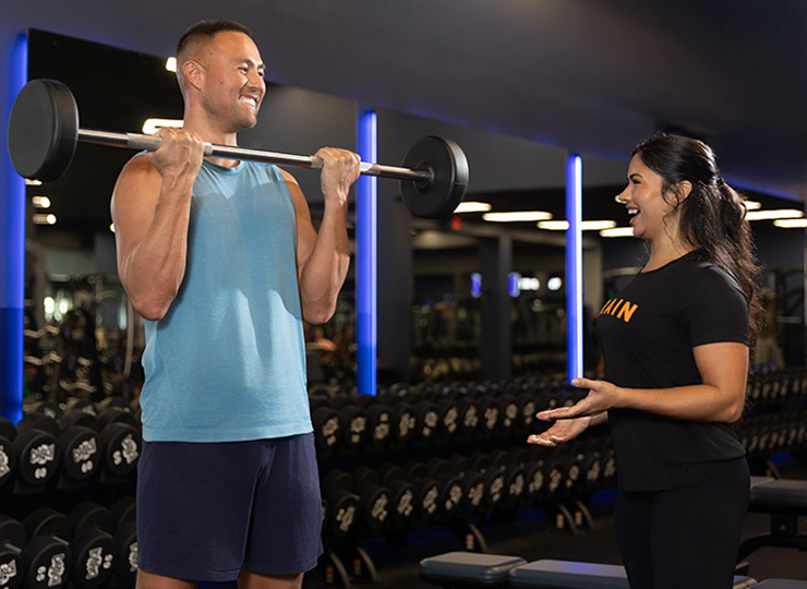 Gym member performing a barbell curl while smiling as a personal trainer coaches and encourages him nearby.