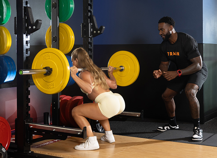 Personal trainer spotting a gym member as she performs a barbell back squat in a squat rack.