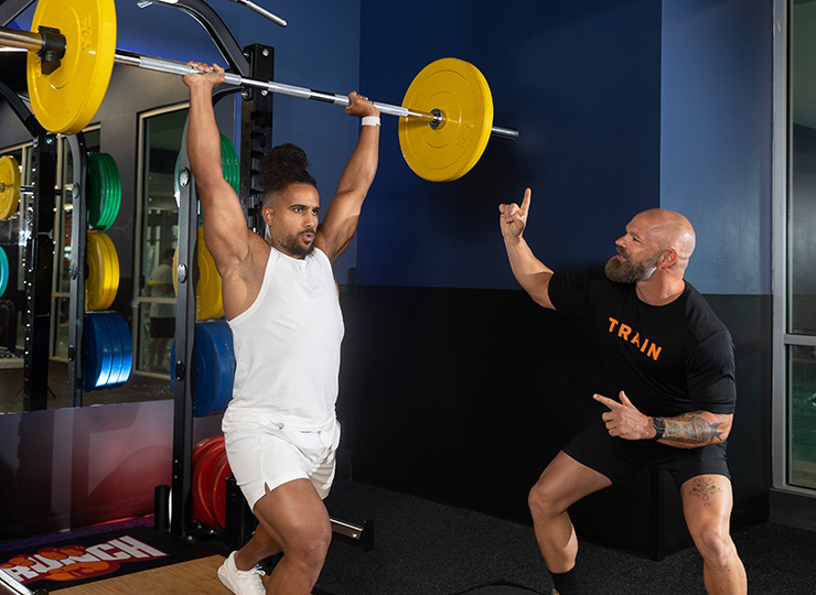 Personal trainer coaching a gym member through a barbell split jerk, with the bar locked out overhead in a split stance.