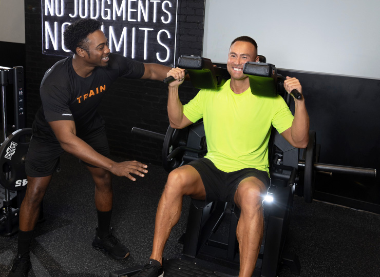 Personal trainer coaching a gym member using a seated shoulder press machine during a strength training session.
