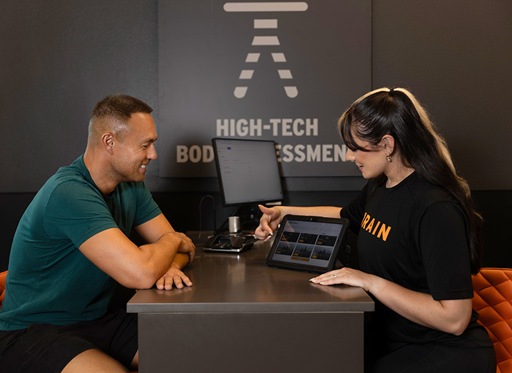 A trainer sits across from a gym member at a consultation desk, pointing to workout information on a tablet beneath a “High-Tech Body Assessment” sign.
