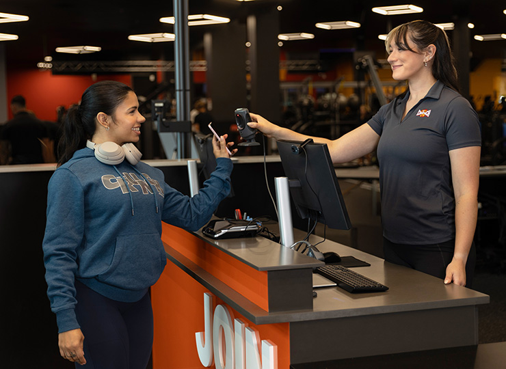 Gym-goer scanning her phone at the front desk while a staff member assists her at check-in.