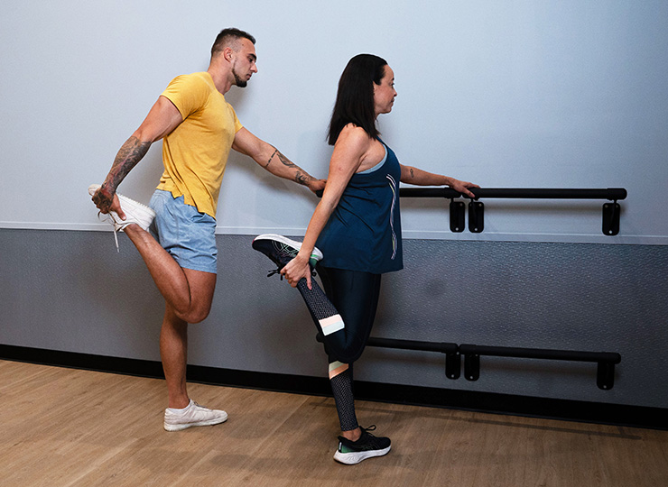 Two people holding onto a barre while doing standing quad stretches in a studio or stretching area.