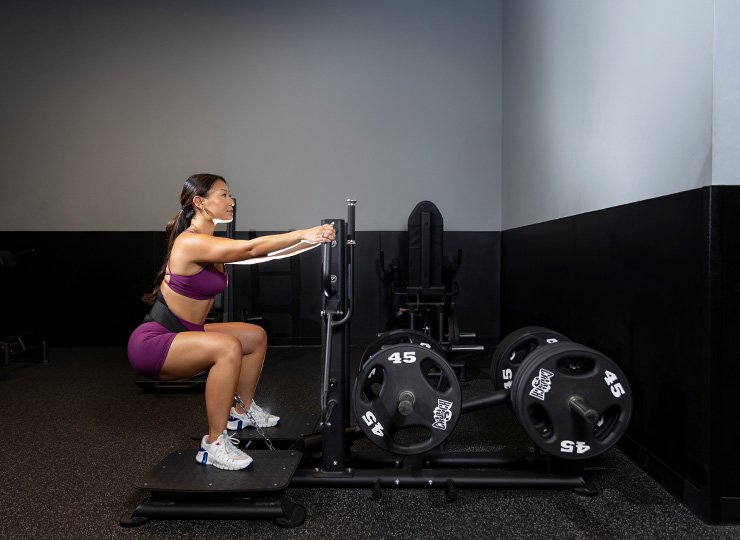 Woman in a purple workout set performing a belt squat on a plate-loaded squat machine in the gym.