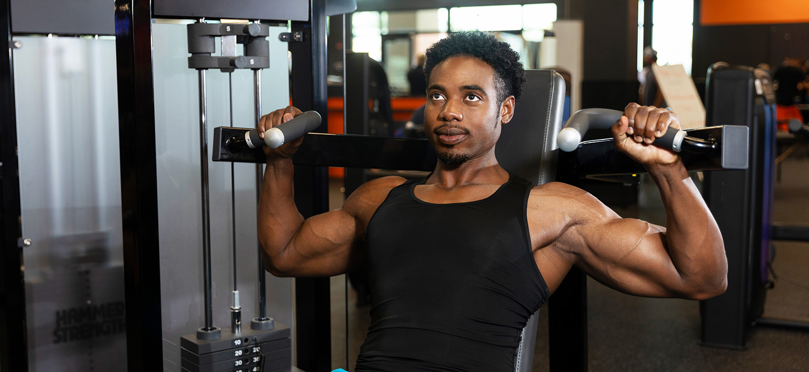 Man using a seated shoulder press machine at the gym, pressing the handles overhead while wearing a black tank top.