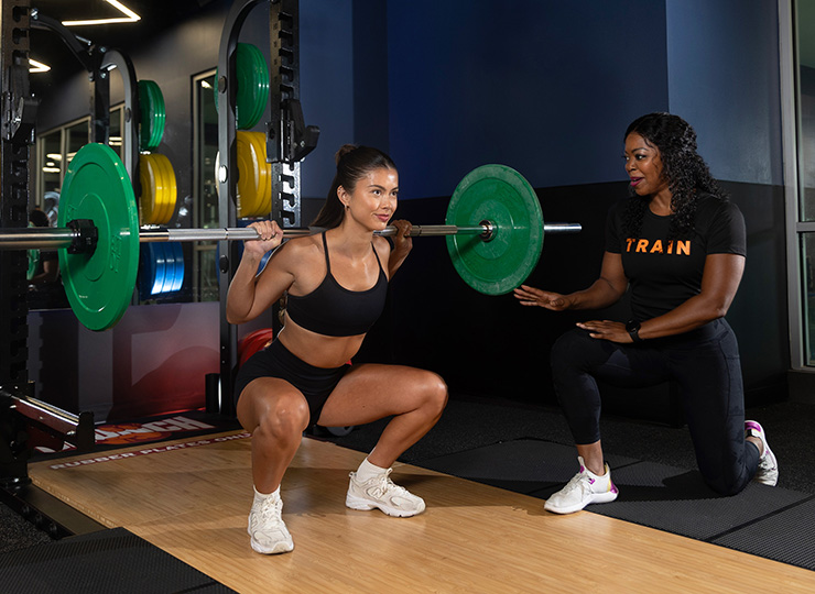 Woman doing a back squat while a trainer coaches beside her.