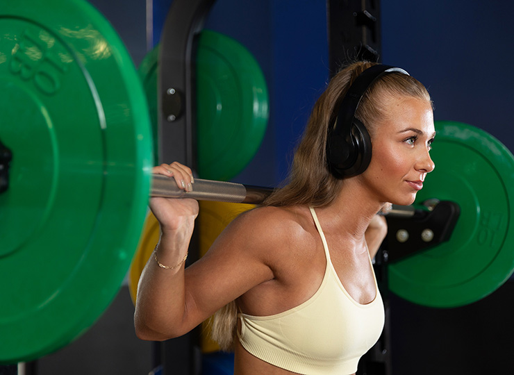 Woman wearing headphones doing a barbell back squat in the gym.