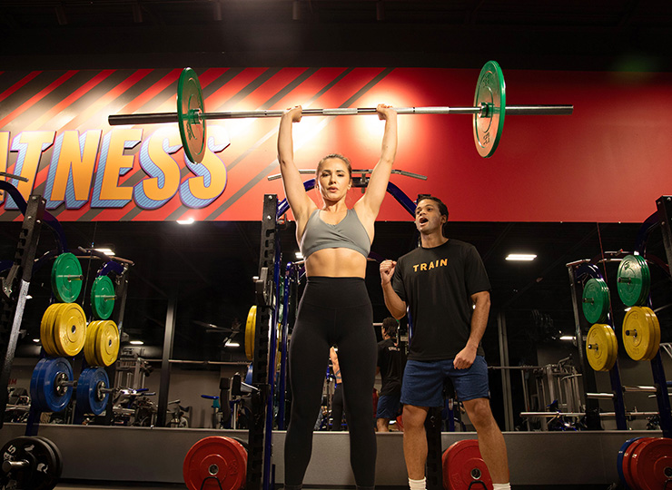 Woman lifting barbell with the help of a personal trainer