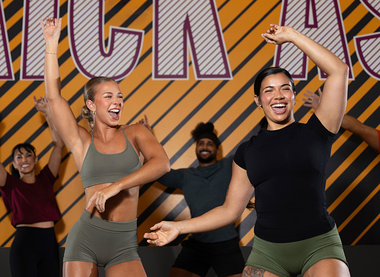 A group fitness class is mid-dance, with two women in the foreground smiling and raising their arms while other participants move behind them against a bold orange-and-black Crunch wall.