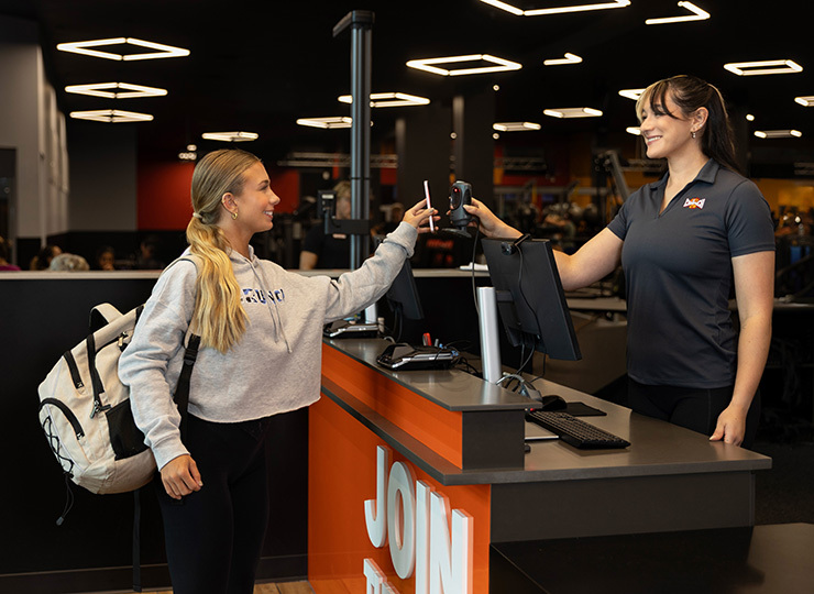 A smiling gym member with a backpack scans into the gym at the front desk while a staff member holds out the barcode scanner.